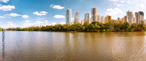 Calm lake in the city of Londrina, Brazil, surrounded by vegetation, with imposing buildings in the background contrasting with nature.