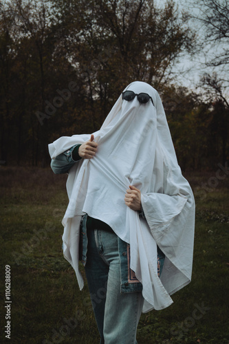 Quadro su tela A person in a white ghost costume with sunglasses poses playfully in a forested area, surrounded by trees with autumn leaves and a moody sky, creating a whimsical yet eerie atmosphere