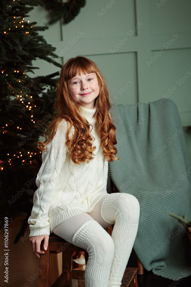 Happy young girl sitting by the Christmas tree in the cozy living room of the house and smiling. Happy winter holidays concept Happy young girl sitting by the Christmas tree in the cozy living room of the house and smiling. Happy winter holidays concept