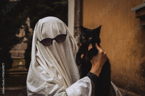 A person wearing a white sheet as a ghost costume and sunglasses gently holds a black cat, both gazing at each other, set against a warm, muted background.