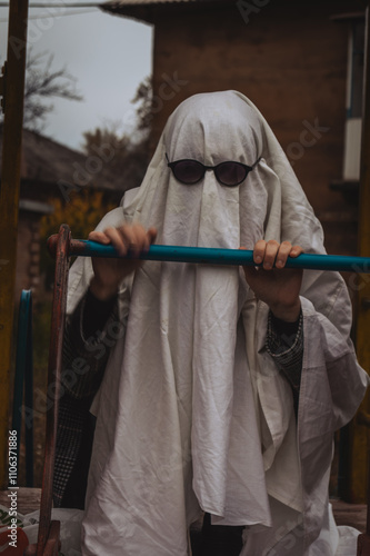 Foto A playful scene shows a ghostly figure draped in a white sheet with sunglasses, seated on a playground slide, evoking humor amidst the mundane backdrop of a residential building