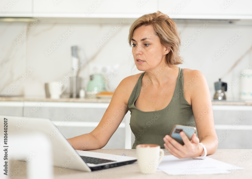 Business-like middle-aged lady working distantly using notebook in cozy tidy kitchen Business-like middle-aged lady working distantly using notebook in cozy tidy kitchen