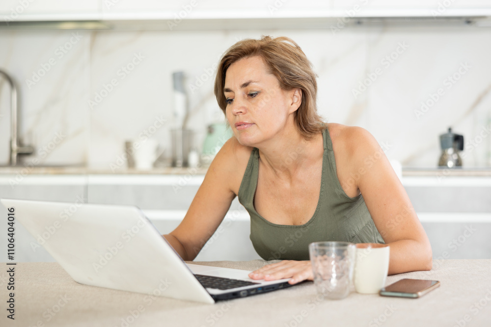 Busy middle-aged woman working diligently on laptop in the kitchen with white interior Busy middle-aged woman working diligently on laptop in the kitchen with white interior