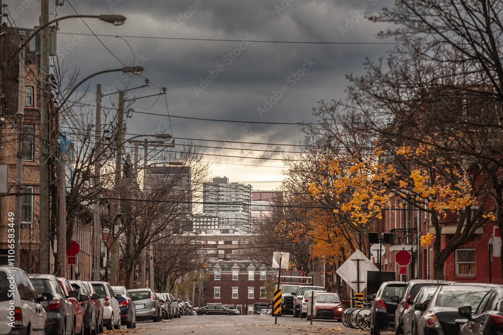 Obraz premium A residential street in Le Plateau, Montreal, Quebec, Canada, in North America during autumn, featuring parked cars, brick houses, and vibrant foliage under a cloudy sky.