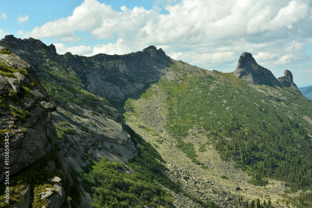 Obraz premium A steep slope of a high mountain with a narrow ridge in a picturesque valley under a summer cloudy sky.