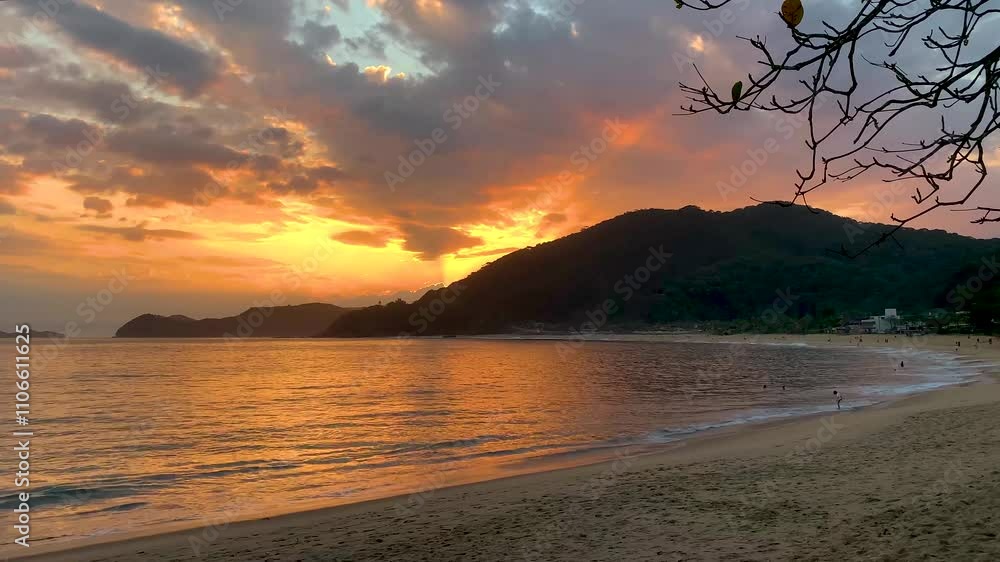 Pôr do sol no mar com o céu dourado, a praia com poucas pessoas e com o sol já atrás das montanhas cobertas com vegetação de Mata Atlântica.