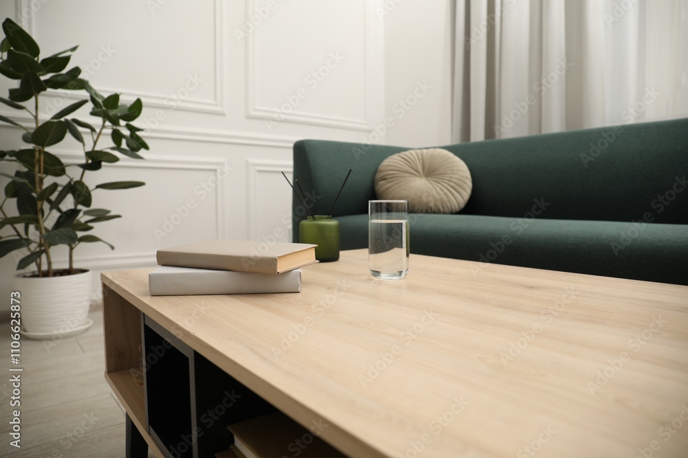 Fototapeta premium Books, glass of water and reed diffuser on wooden coffee table near sofa in living room, closeup