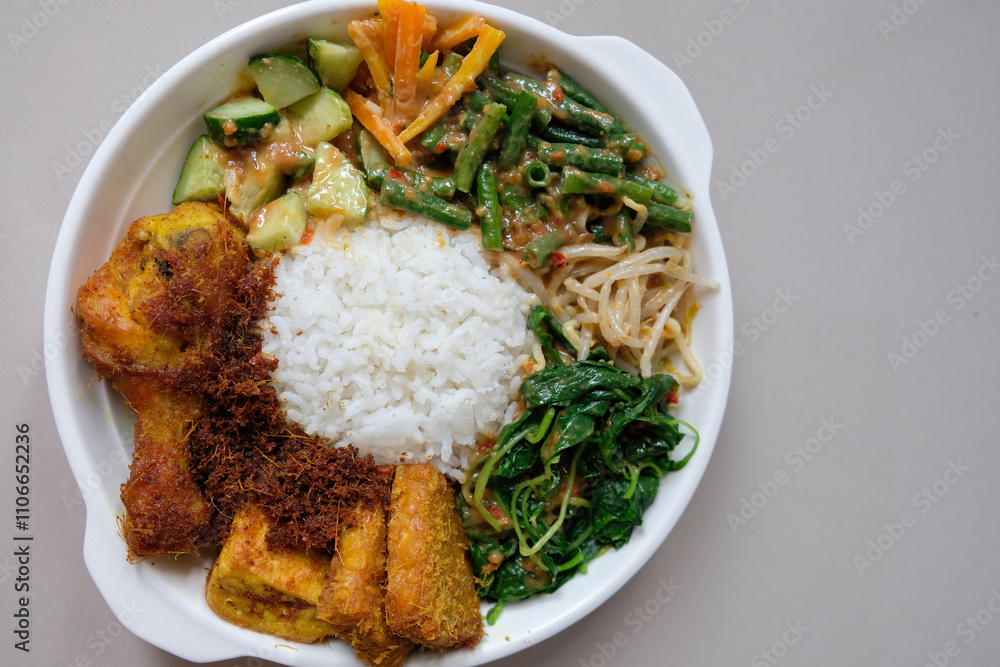 An Indonesian homemade food, rice with pecel, tempeh, tofu and ayam goreng lengkuas or galangal fried chicken on a plate. Top view, copy space. An Indonesian homemade food, rice with pecel, tempeh, tofu and ayam goreng lengkuas or galangal fried chicken on a plate. Top view, copy space.