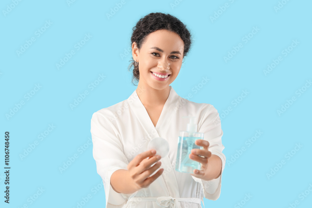 Happy young African-American woman with bottle of micellar water and cotton pad on blue background Happy young African-American woman with bottle of micellar water and cotton pad on blue background