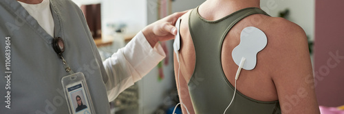 Doctor attaching medical electrodes on patient's back for receiving treatment in clinic setting. Medical professional wearing identification badge and ensuring proper placement of electrodes