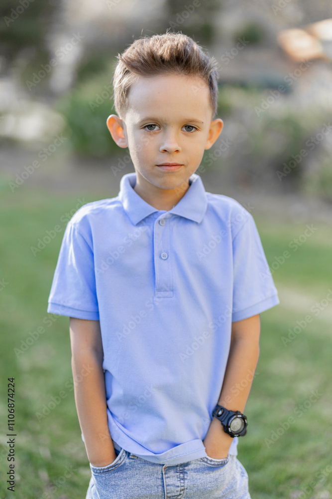A young boy is standing confidently with his hands resting in the pockets of his light blue polo shirt, set against a charmingly soft outdoor background that beautifully enhances his calm demeanor