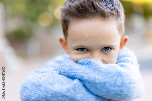 A charming and delightful young boy is wrapped snugly in a soft and cozy blue sweater, gleefully looking playfully at the camera while exhibiting a warm and inviting smile that radiates happiness