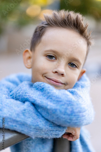 A joyful young boy in a cozy blue sweater beams against a softly blurred, colorful outdoor backdrop, capturing the pure essence of childhood happiness and innocence we all cherish