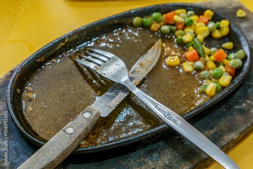 A minimalist food photography shot of a stainless steel steak knife and fork placed diagonally on a hot plate, surrounded by the remains of a gourmet meal. photo