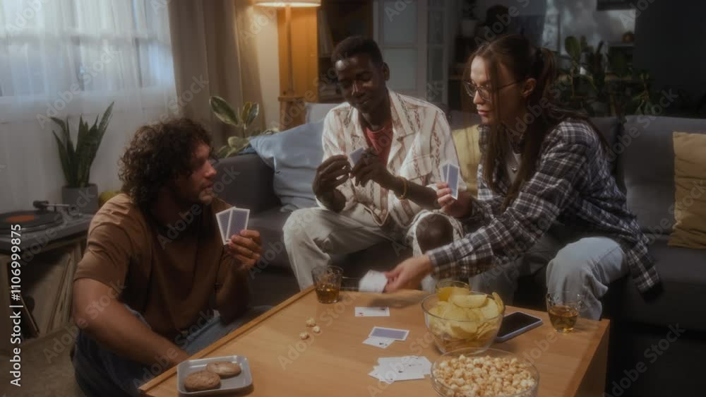 Group of three young male and female friends having fun playing card game in living room at home Group of three young male and female friends having fun playing card game in living room at home
