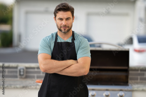 male chef in an apron stands by the grill outside and looks at camera. man is cooking on a grill. chef man at barbecue and grill . chef at barbecue and grill. Man grilling food on barbecue in backyard