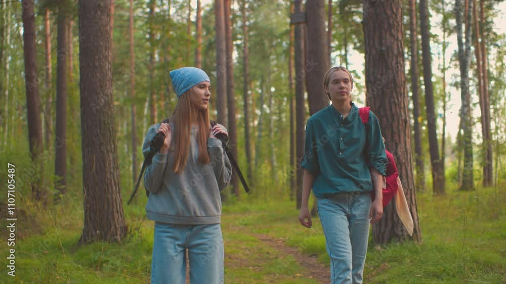 Two ladies walk along forest trail surrounded by lush greenery, one playfully swings her black bag over her shoulder, while the other watches with a warm smile Two ladies walk along forest trail surrounded by lush greenery, one playfully swings her black bag over her shoulder, while the other watches with a warm smile