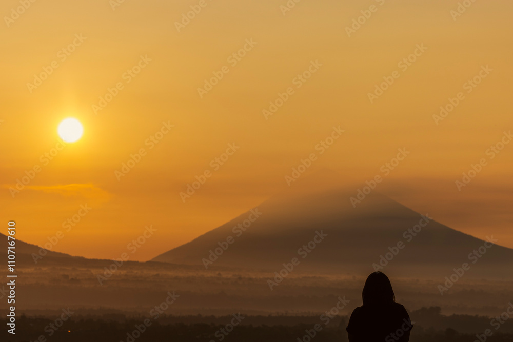 A solitary figure gazes at the sunrise over Mount Agung, silhouetted against a golden sky as morning light filters through layers of mist and softens the distant volcanic peak. A solitary figure gazes at the sunrise over Mount Agung, silhouetted against a golden sky as morning light filters through layers of mist and softens the distant volcanic peak.