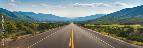 A long, straight road stretches towards the horizon, flanked by lush mountains and a blue sky.