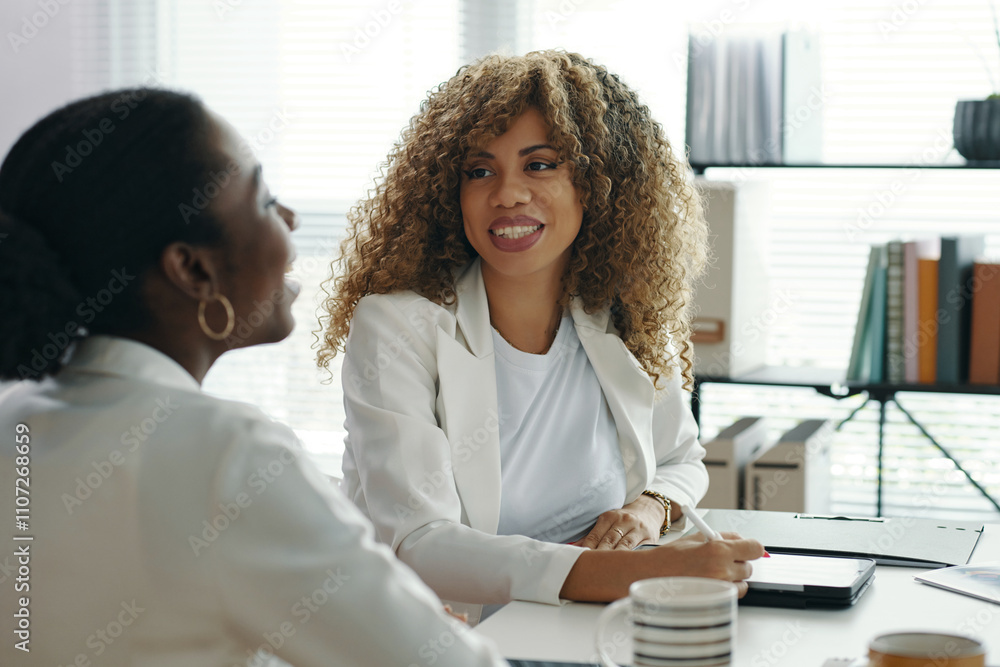 Two businesswomen discussing work in modern office setting. One smiling, facing camera, with curly hair, wearing white. Bright, professional ambiance with bookshelves and files in background Two businesswomen discussing work in modern office setting. One smiling, facing camera, with curly hair, wearing white. Bright, professional ambiance with bookshelves and files in background