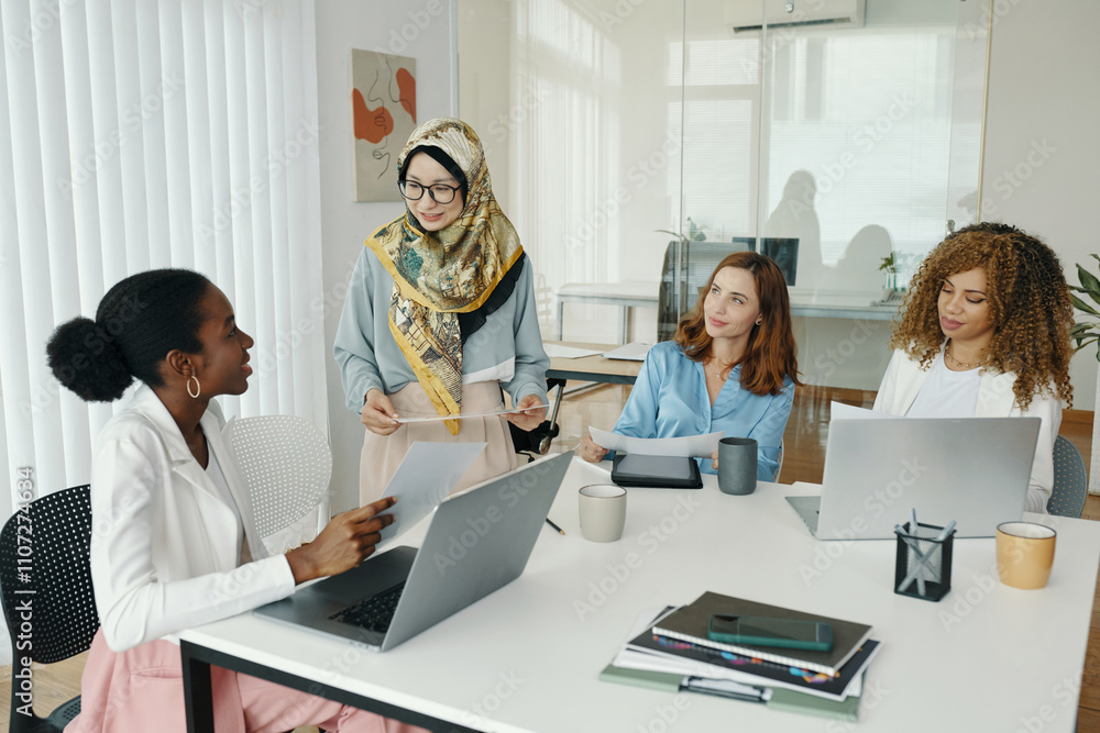 Diverse group of women discussing work while seated around table in modern office. Collaboration and teamwork are evident. Various devices and documents are spread across the table Diverse group of women discussing work while seated around table in modern office. Collaboration and teamwork are evident. Various devices and documents are spread across the table