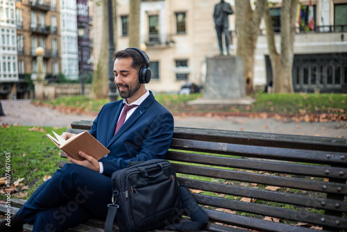 Businessman listening music with headphones and reading book on bench