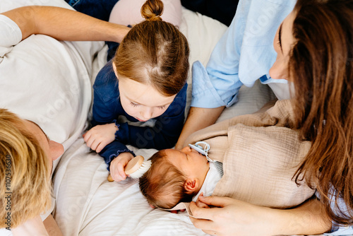 Top view of loving family bonding with newborn baby. Little sister brushes baby s hair while sits on comfortable bed. Parents, older sister, and infant boy on cozy bedroom interior. photo