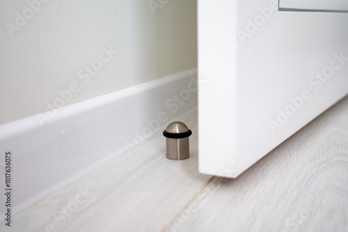 Door stopper on a light wooden floor next to a white door and wall skirting, providing protection from door impact photo