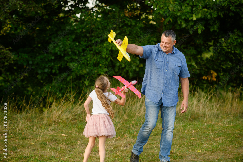 Fototapeta premium Happy girl and dad having fun outdoors, playing with toy airplane, running with it, in park on grass, smiling happily. Childhood and dream concept, father and child games. International Father's Day