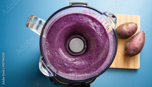 A blender contains a vibrant purple smoothie or puree with sweet potatoes visible on a cutting board beside it, viewed from above. photo