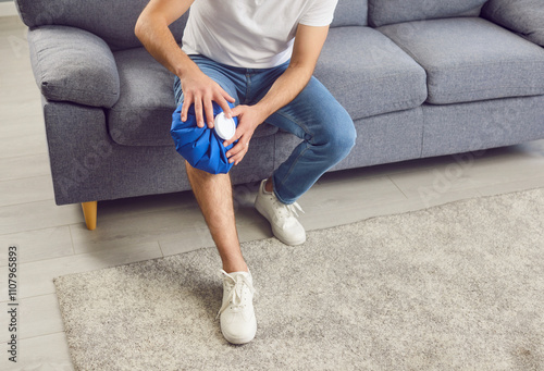 Man, sitting on sofa at home and using an ice pack on leg to relieve pain after an injury. The cold treatment helps soothe the ache in hurt knee, providing comfort and reducing inflammation. photo