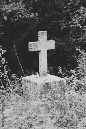 Old stone cross in overgrown graveyard. Weathered memorial tombstone surrounded by wild plants. Atmospheric black and white cemetery image for All Saints' Day or Halloween design photo