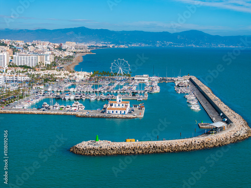 The image captures Benalmadena's marina with yachts, a Ferris wheel, and a curved breakwater. Distant mountains and town buildings are visible under a clear sky. photo