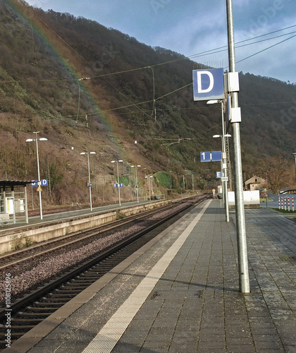 A small railway station in the mountains. There is a colorful rainbow in the background