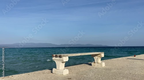 A bench on the shore of the Aegean Sea against the background of blue waves. Turquoise water and a beautiful view. Sunny day by the sea in Greece. High quality 4k video