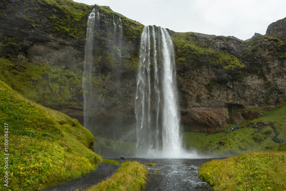 Obraz premium The beautiful Seljalandsfoss waterfall in South Iceland offers a unique perspective, with pathways allowing visitors to admire its beauty from behind the falls. Tourists enjoying Iceland.