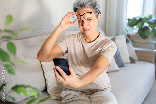 Bilde på lerret A middle-aged woman sits on the couch and lifts her glasses trying to read a message on the phone