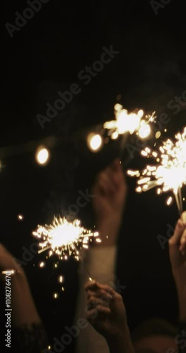 VERTICAL VIDEO: Burning sparklers in hands of group of friends on christmas. People waving new year lights in air on black background. Friends at party