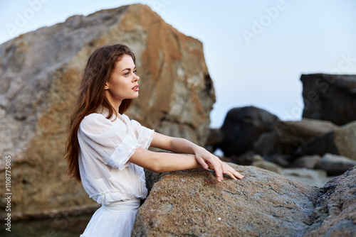 Serene woman in white dress poses confidently on rocky ocean shore with hands on hips photo