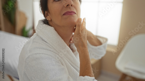 Woman applying cream in a wellness spa room, wearing a white robe and looking upwards, reflecting mature beauty in a serene indoor setting.