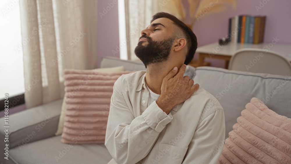 Young hispanic man with a beard sitting in a cozy living room at home rubbing his neck in discomfort Young hispanic man with a beard sitting in a cozy living room at home rubbing his neck in discomfort