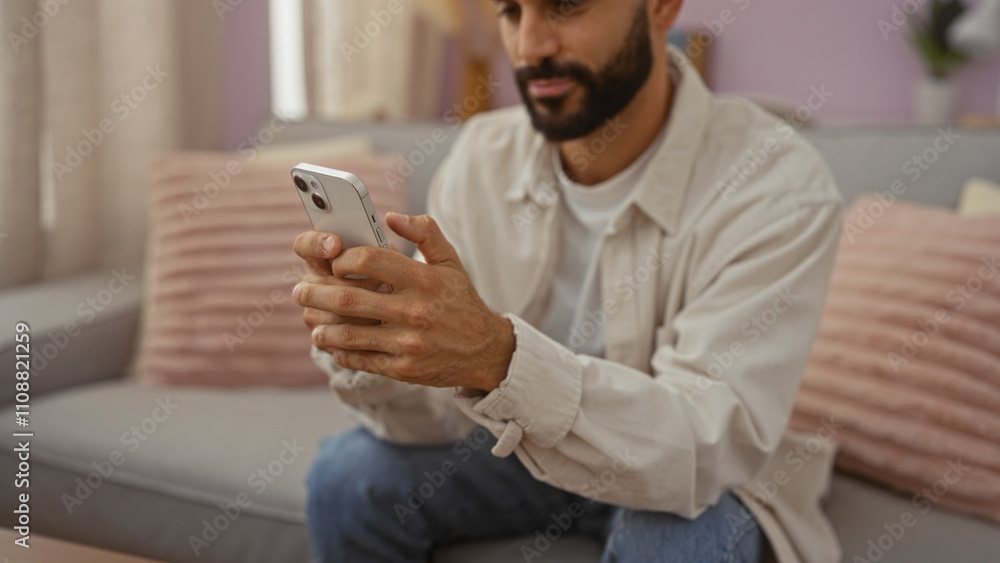 Young hispanic man with beard using a smartphone while sitting in a cozy living room with pink pillows and beige curtains Young hispanic man with beard using a smartphone while sitting in a cozy living room with pink pillows and beige curtains