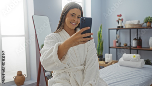 Woman taking selfie at spa in wellness center indoor wearing bathrobe with smile in serene beauty salon background