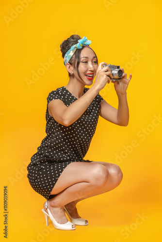 Emotional Asian girl dressed in pin-up style with vintage camera in her hands doing photo shoot on bright yellow background. Isolated.