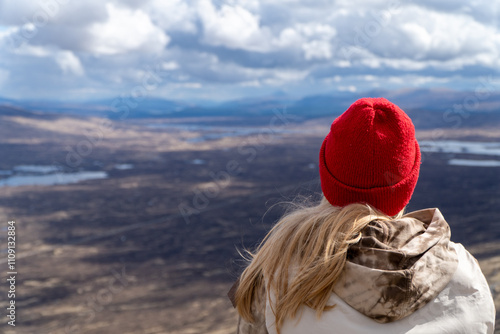 This image captures a person wearing a vibrant red beanie and a tie-dye hoodie, gazing over a vast, rugged landscape with water bodies, hills, and dramatic skies in the distance.