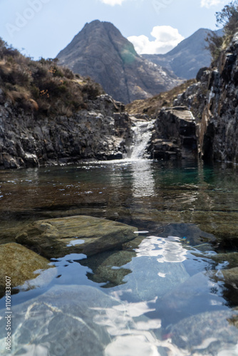 This image captures a serene mountain pool with crystal-clear water and a small waterfall framed by rocky terrain. In the background, towering, rugged mountains rise against a partly cloudy sky.