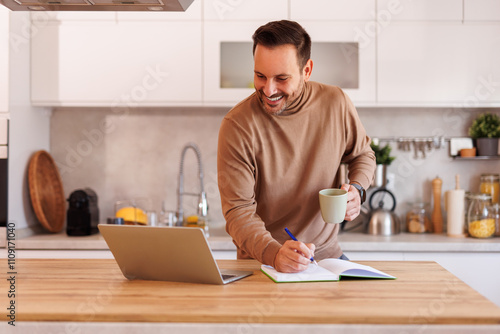 Mid adult man holding coffee mug and writing in note pad while working over laptop at table in kitchen