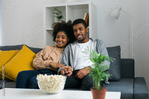 African American couple relaxes on a cozy sofa, sharing a joyful moment while watching a show with a bowl of popcorn. Essence of quality time and comfort in a stylish modern living room photo