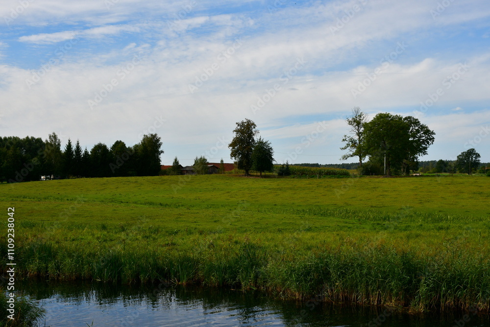 A view of a shallow yet vast pond or lake flowing next to vast fields covered with herbs, reeds, and various crops located next to some tiny forests and moors spotted on a cloudy summer day in Poland A view of a shallow yet vast pond or lake flowing next to vast fields covered with herbs, reeds, and various crops located next to some tiny forests and moors spotted on a cloudy summer day in Poland