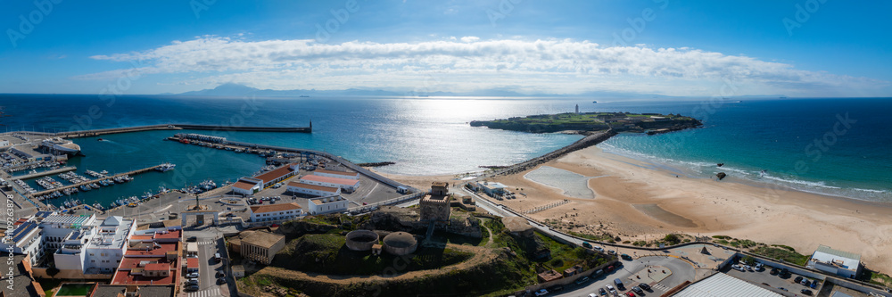 Obraz premium Tarifa's coastal landscape features Isla de las Palomas, a lighthouse, bustling harbor, sandy beaches, kite surfers, and the distant African coast.